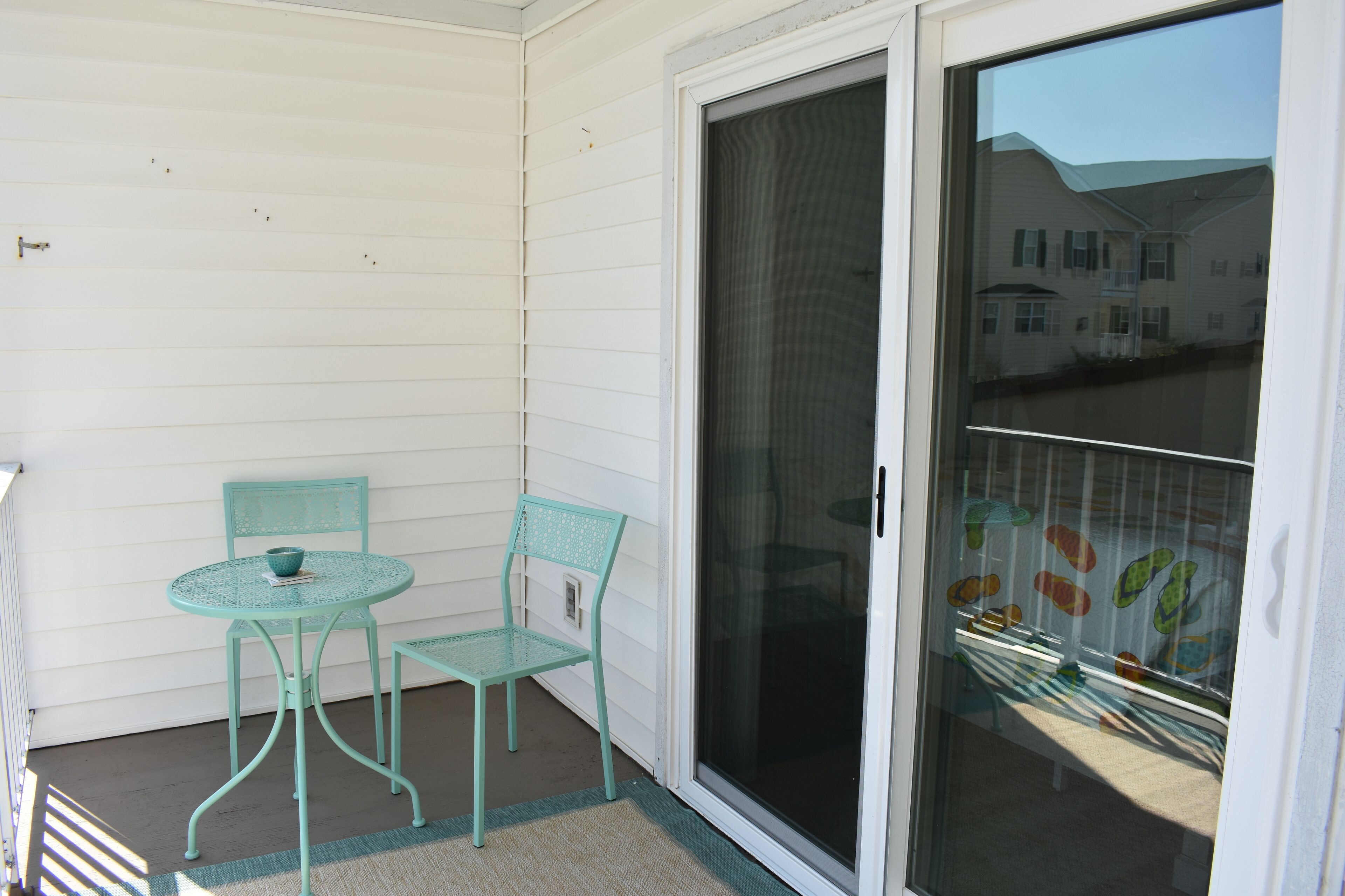 Ein kleiner Balkon mit zwei türkisfarbenen Metallstühlen und einem Tisch, eine Schiebetür aus Glas sowie Blick auf ein Wohngebiet.