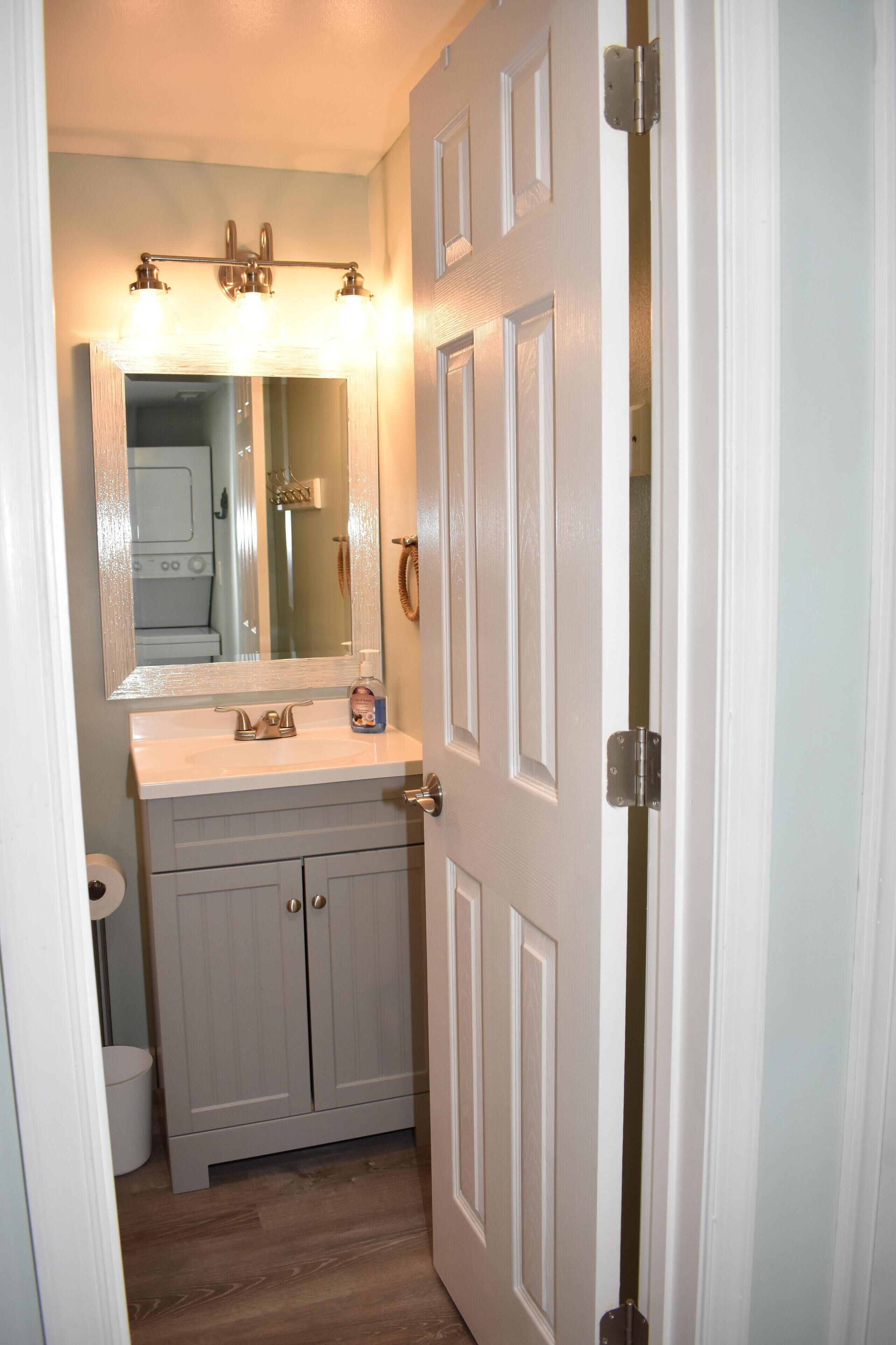 Une salle de bain avec un meuble-lavabo blanc, un miroir et une porte dotée d’un interrupteur.