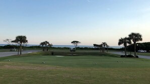 A golf course with a clear view of the ocean, palm trees, and a white truck parked on the side. - Sand Castle, 9 bedroom,6 1/2 bath,sleep 26,  pool, hottub, elevator, beach close (Oak Island)