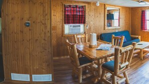 Interior de una cabaña de madera con una mesa de comedor, sillas, un sofá y una ventana con cortinas rojas.