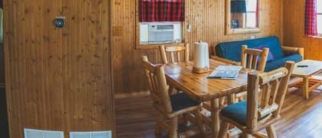 A wooden interior with a dining table, chairs, a sofa, and a window with a red and black checkered curtain.
