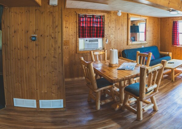 A wooden interior with a dining table, chairs, a sofa, and a window with a red and black checkered curtain. - 2BR Lakefront Cabin w/ Private Launch & Dock (Backus)