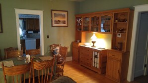A dining area with a wooden table and chairs, a kitchen with wooden cabinets, and a lit table lamp.