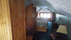 A room with a wooden door, a bed with a patterned bedspread, a nightstand with a lamp, and a small table.