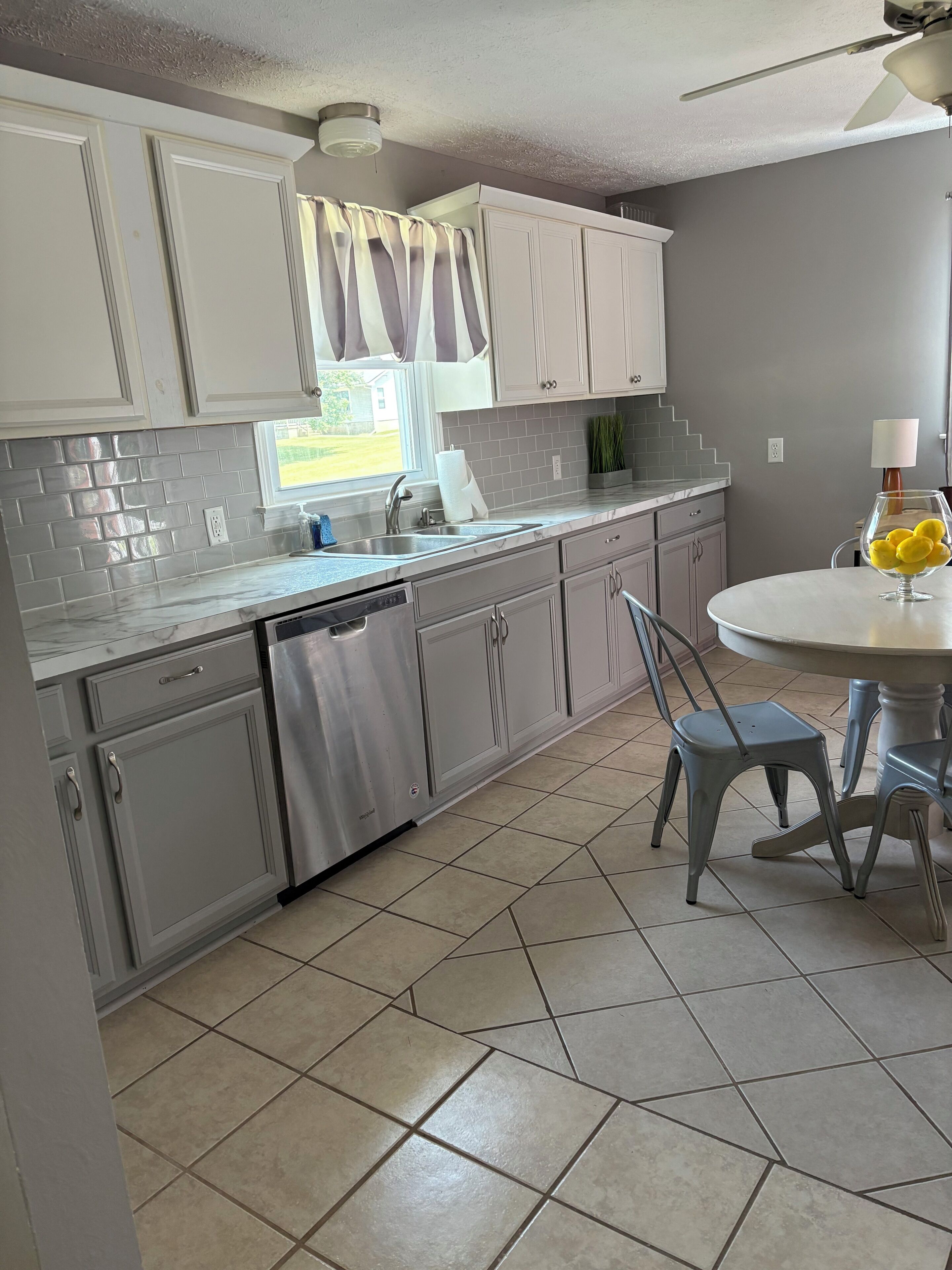A kitchen with white cabinets, a dishwasher, a window with curtains, and a dining area with a table and chairs.