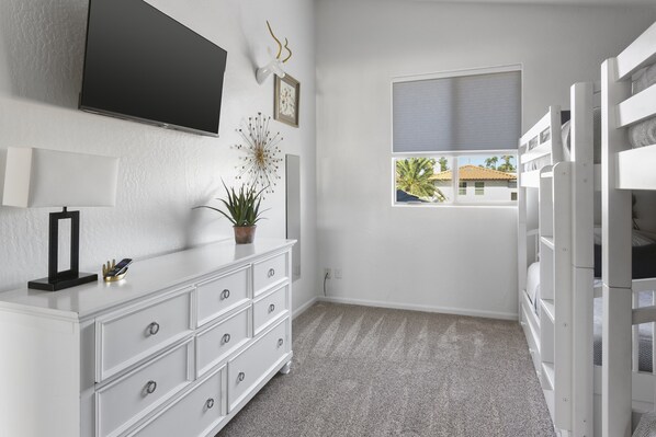 A room with a white dresser, a flat-screen TV mounted on the wall, a potted plant, and a window with a view of palm trees.