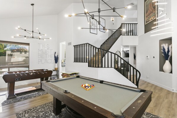 Uma mesa de bilhar com bola branca, uma mesa de futebol de mesa e uma escadaria com guarda-corpos pretos.