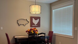 A dining area with a wooden table, four chairs, a vase of flowers, a wall-mounted light fixture, and a window with blinds. - All One Level Basement Unit Large Great Room Kitchen 3 bed 2 baths (Lava Hot Springs)