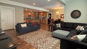 A spacious living room with a large black sofa, a blue sofa, and a patterned rug. There is a kitchen in the background with wooden cabinets and a refrigerator. - All One Level Basement Unit Large Great Room Kitchen 3 bed 2 baths (Lava Hot Springs)