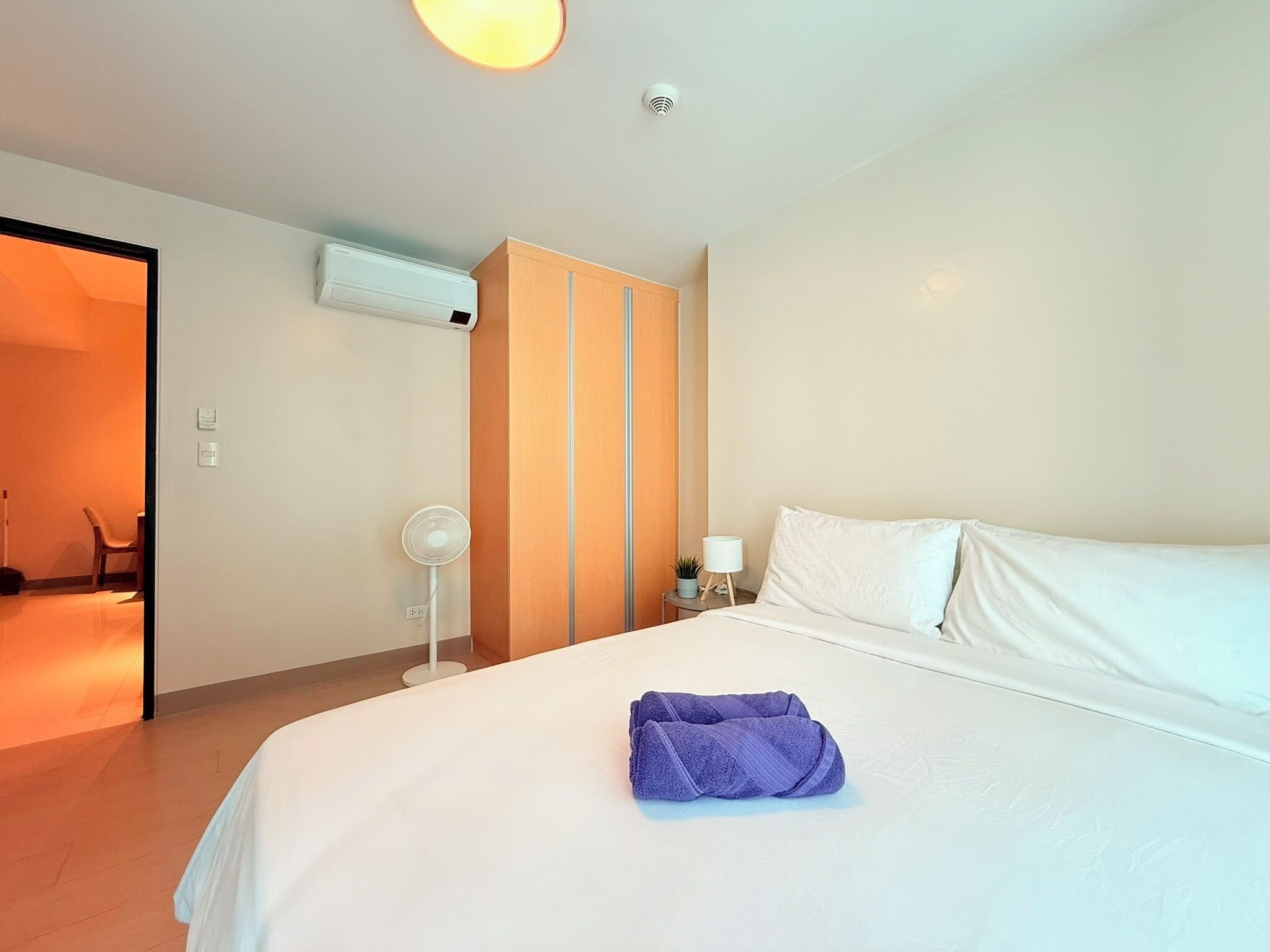 A hotel room with a bed, a nightstand, a lamp, a fan, and an air conditioning unit.