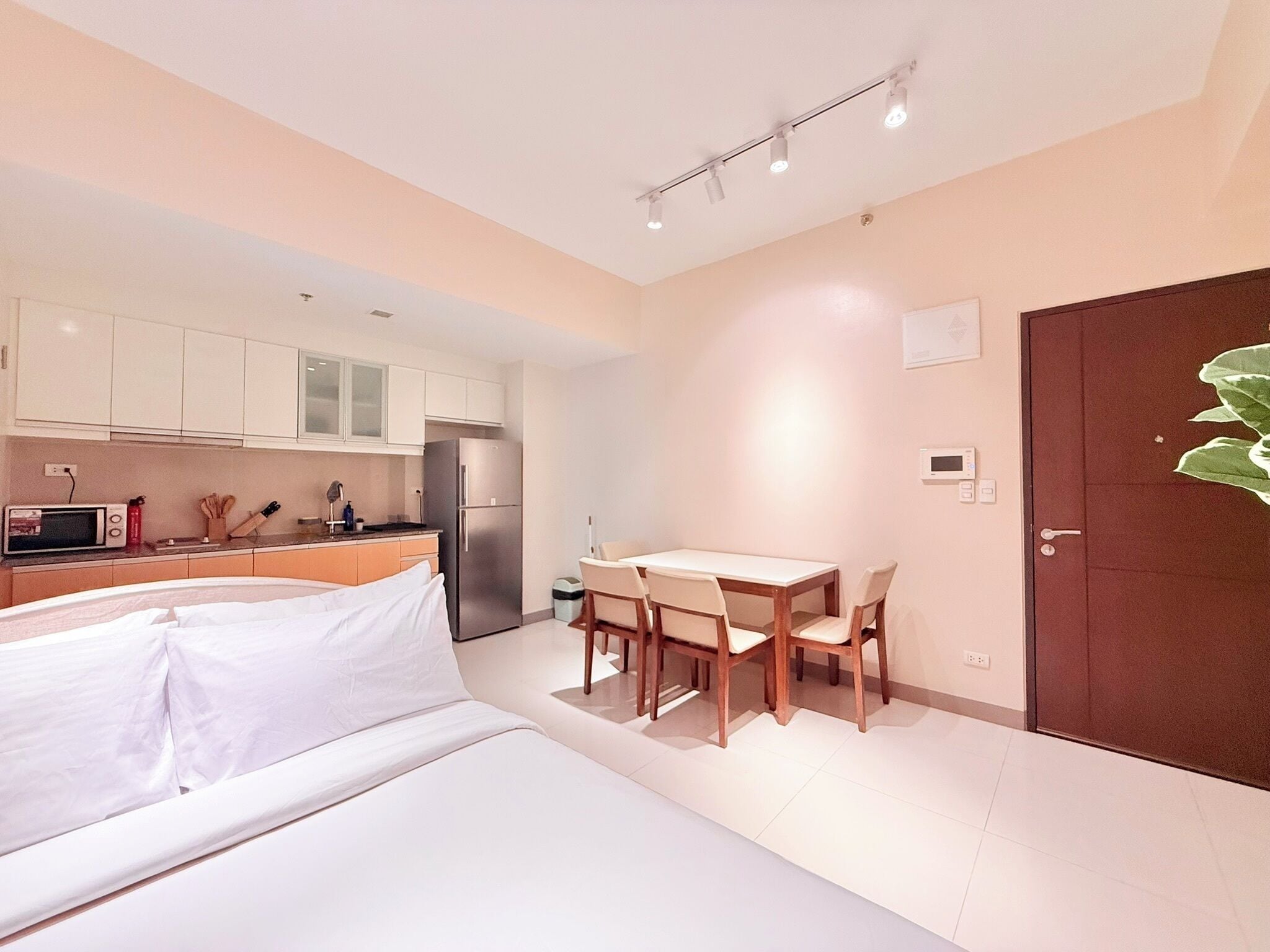 A compact hotel room with a bed, a small kitchenette, a dining table with chairs, and a door with a plant decoration.
