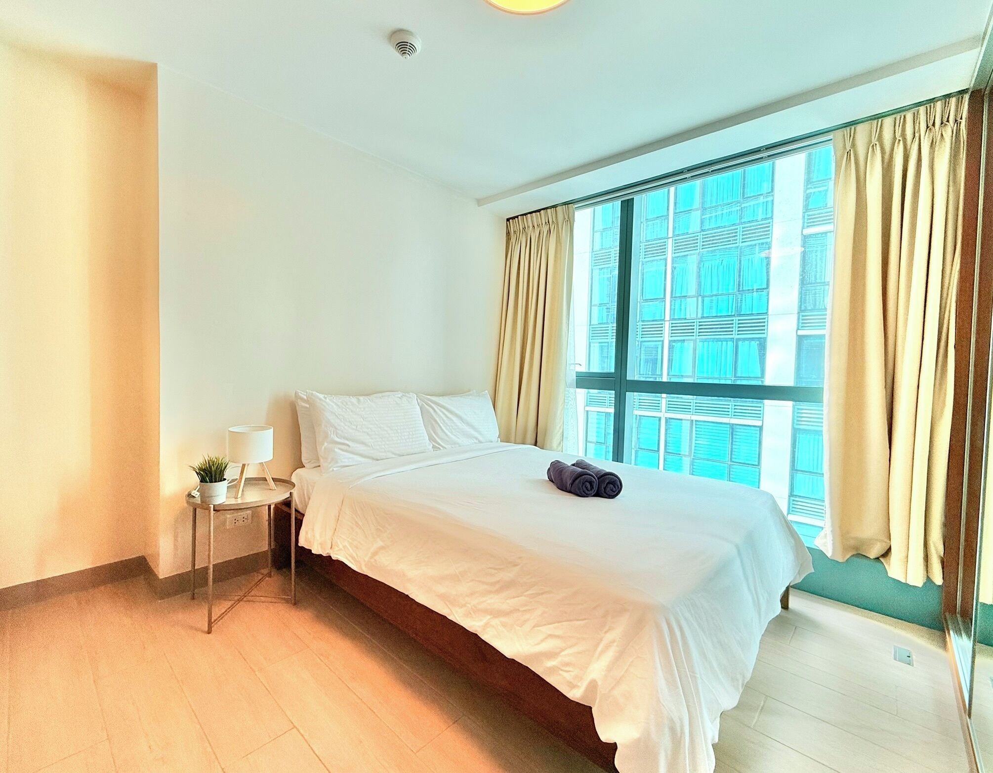 A hotel room with a large bed, a bedside table with a lamp, and a view of a building through the window.