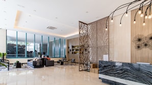 A modern reception area with a marble reception desk, a seating area with sofas and chairs, large windows offering views, and a decorative wall with a light fixture. - One Uptown Residence BGC (Taguig)