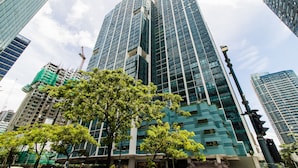 A modern high-rise building with a glass facade, surrounded by other tall structures and trees. - One Uptown Residence BGC (Taguig)