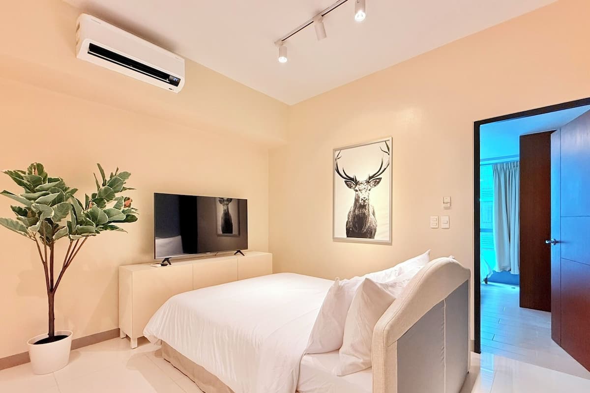 A modern bedroom with a bed, a flat-screen TV, a potted plant, and a deer artwork on the wall.