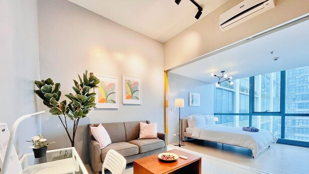 A modern hotel room with a large bed, a sofa, a coffee table, and a view of the city through large windows. - One Uptown Residence BGC (Taguig)