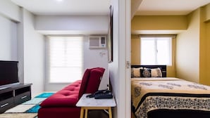 A hotel room with a bed, a red sofa, a small table with a phone, a TV, and a window with blinds.