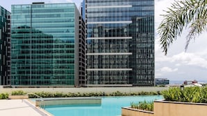 A modern building with a glass facade, a rooftop pool, and surrounding greenery.