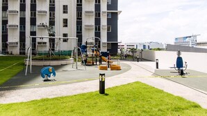A playground with various equipment, a blue and orange slide, and a climbing structure, surrounded by a grassy area and a building with multiple balconies in the background.