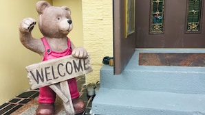 A bear statue holding a 'WELCOME' sign at the entrance of a building with a yellow wall and a brown door. - Coffee Bear (Hengchun)