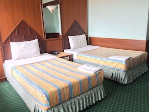 A hotel room with two beds, each with a striped bedspread and white pillows. There is a wooden headboard, a small bedside table with a lamp, and a large mirror on the wall. - Grand Thara Hotel (Surat Thani)