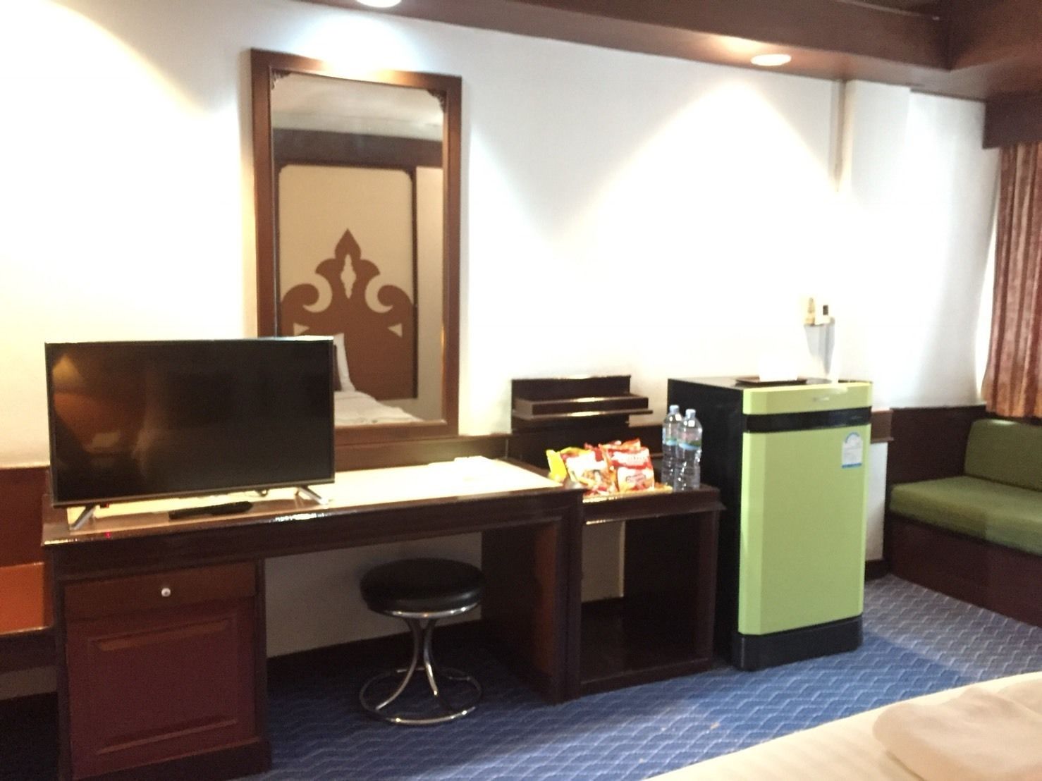 A hotel room with a bed, a desk with a chair, a television, a minibar, and a green sofa.