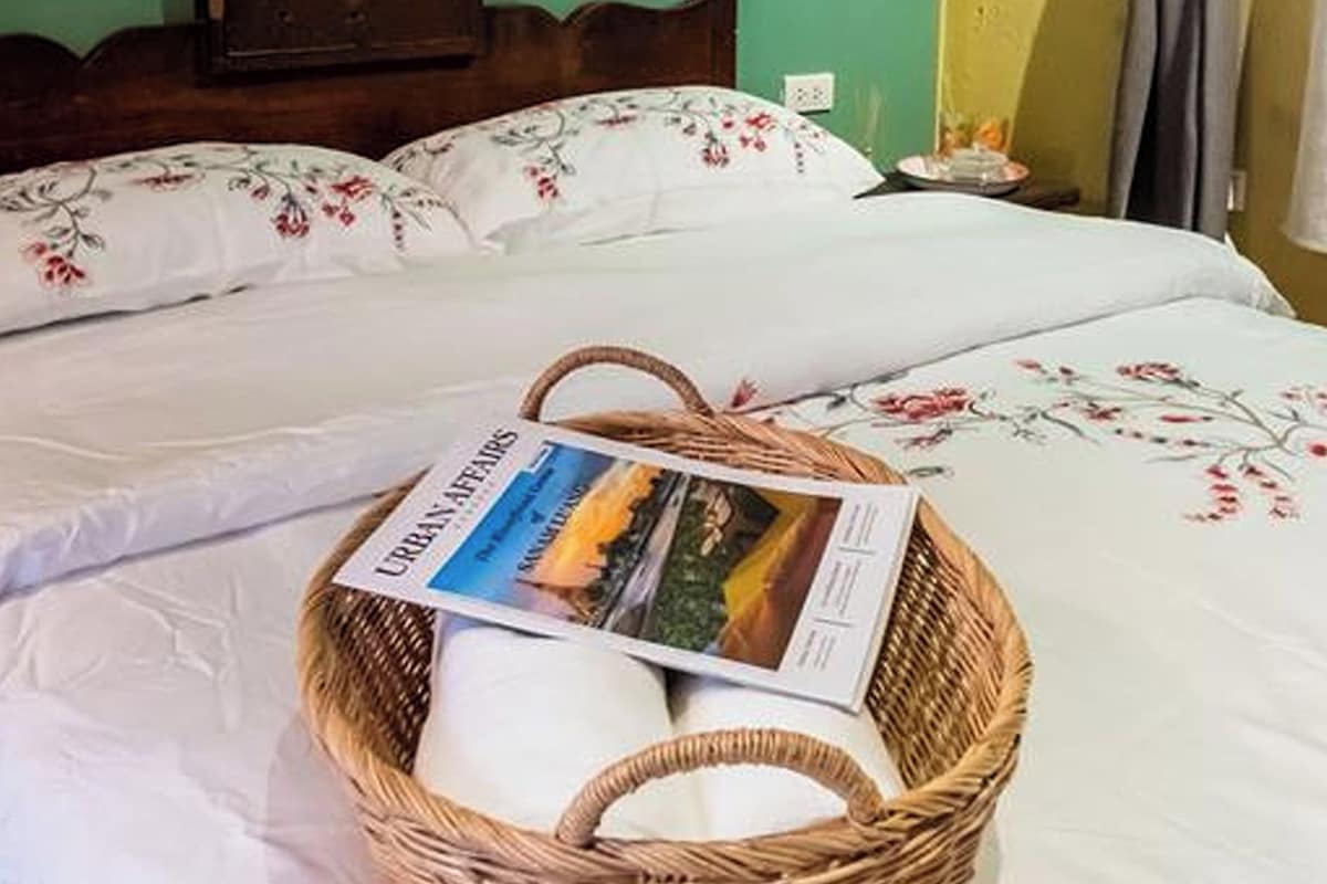 A neatly made bed with floral bedding, a wicker basket with a magazine, a wooden headboard, and a wall-mounted painting.