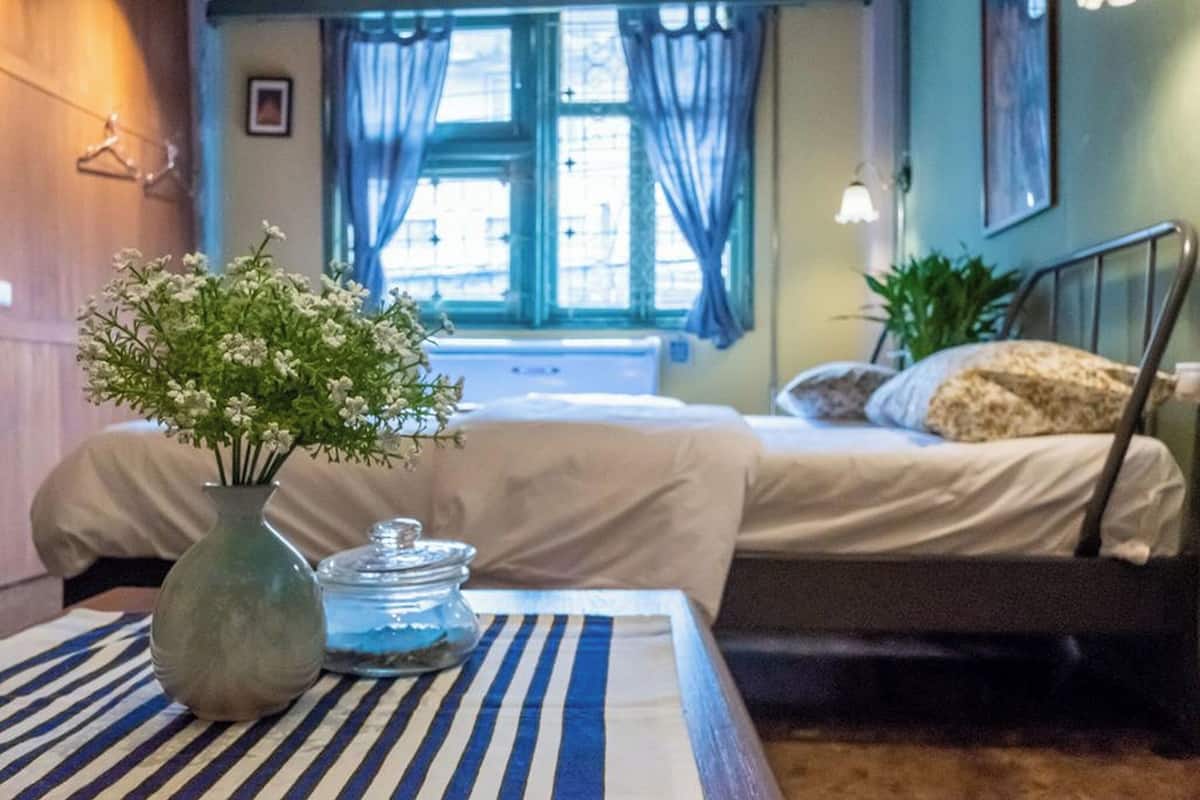A room with a bed, a bedside table with a vase and a jar, a window with curtains, and a wall-mounted lamp.