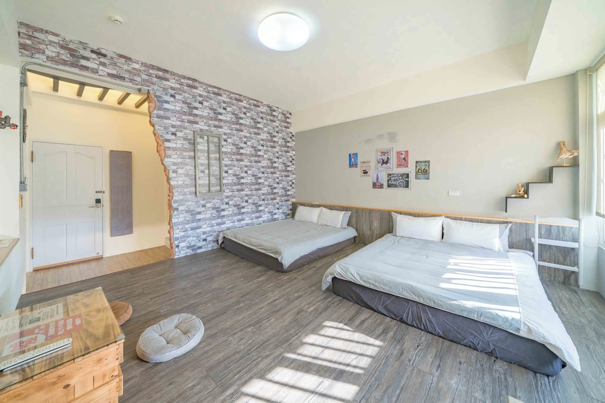 Quarto com duas camas, parede de tijolos, piso de madeira e porta que dá para outro cômodo.