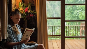 A person reading a book in a room with a wooden deck visible through a large window. - Our Jungle House (Phanom)