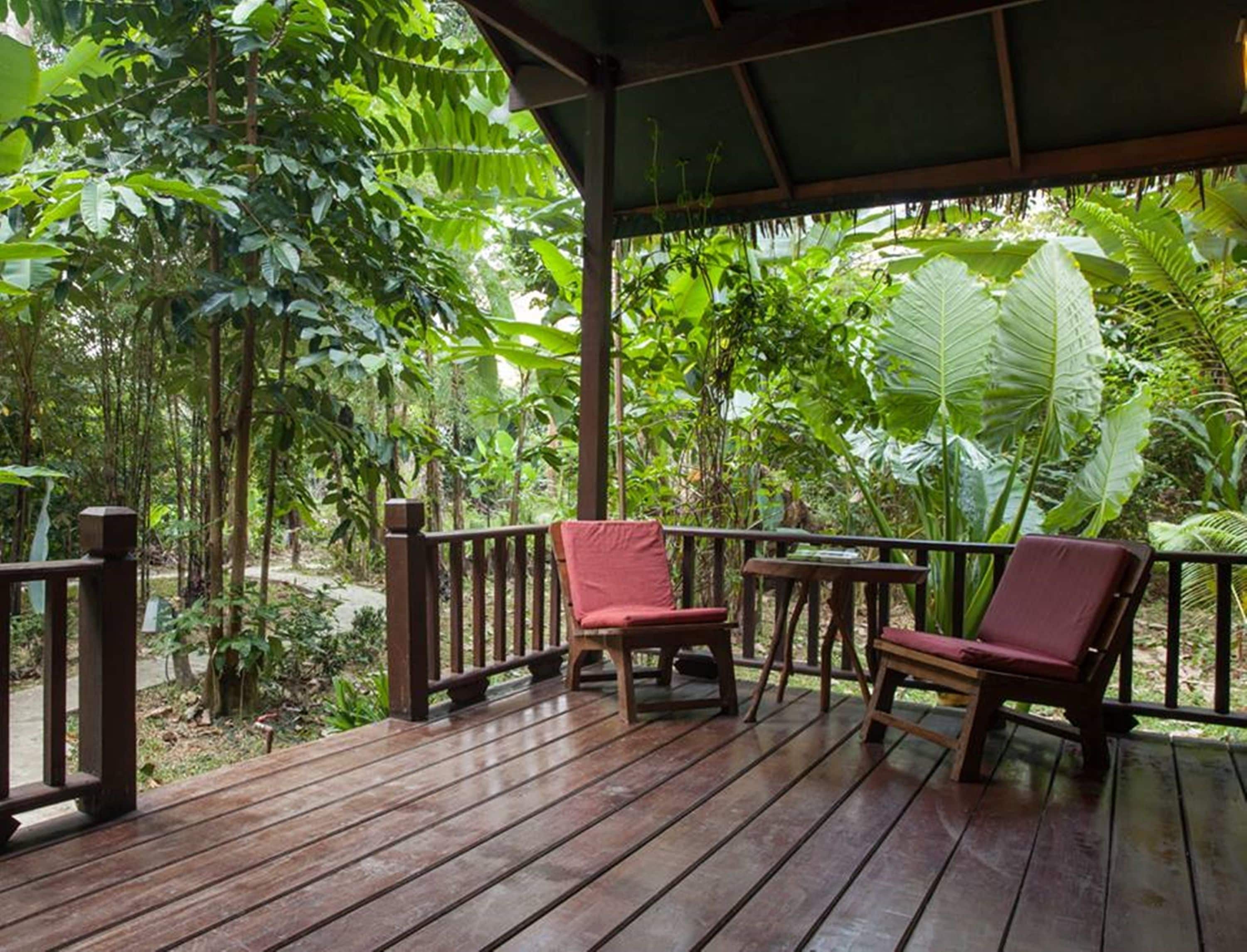 A wooden deck with two red chairs and a table, surrounded by lush tropical vegetation.