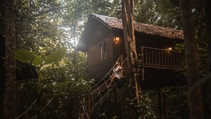 Een houten boomhut met een rieten dak, omgeven door weelderige begroeiing.
