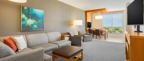 A modern hotel room with a grey sofa set, a wooden coffee table, a flat-screen TV, and a large window offering a city view.