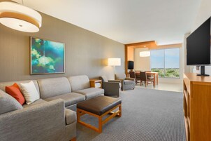 Quarto de hotel moderno com sofá cinzento, mesa de centro de madeira, televisão de tela plana e uma janela grande com vista para a cidade.