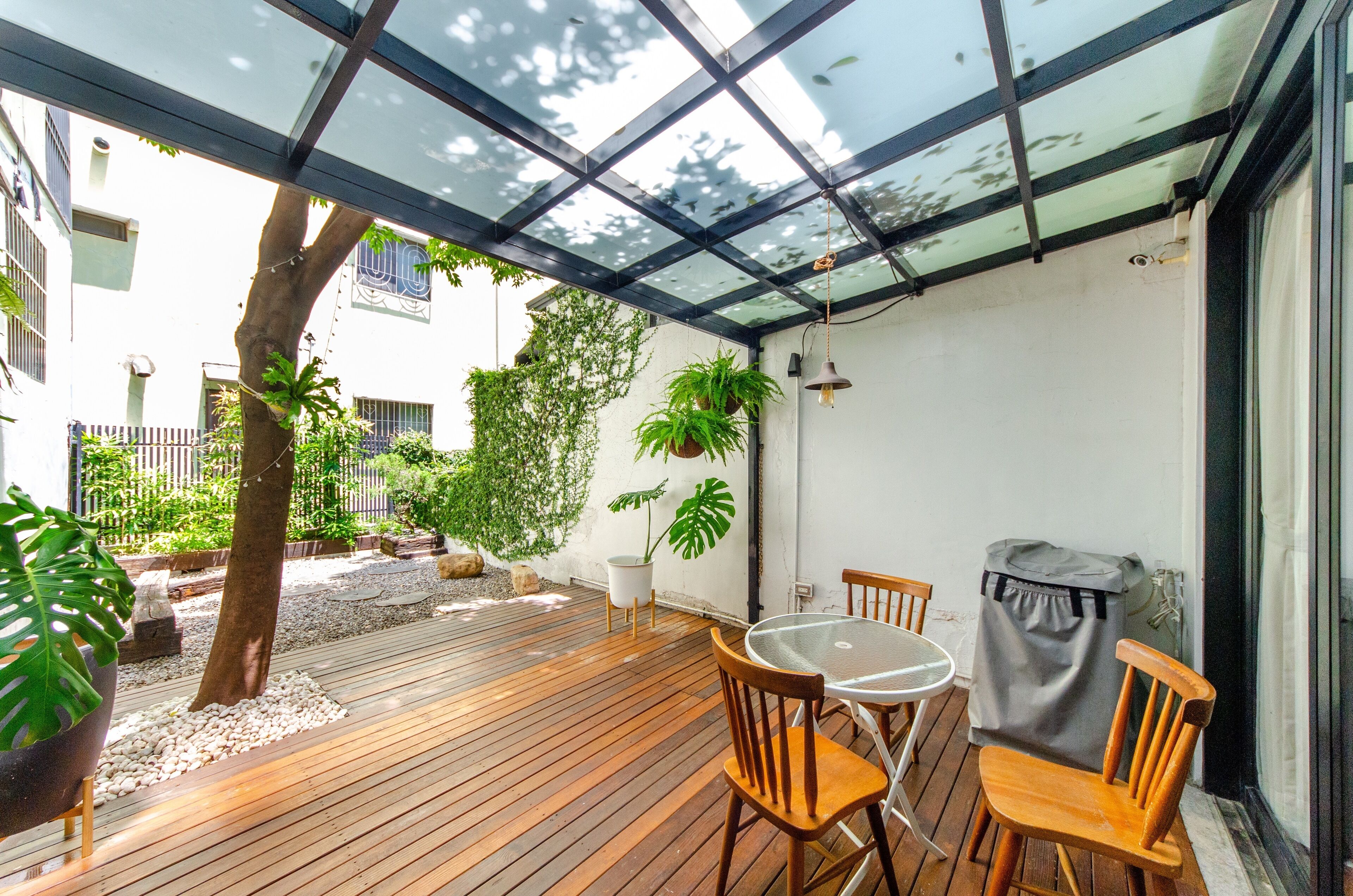 A wooden deck with a glass ceiling, surrounded by potted plants and a small table with chairs.