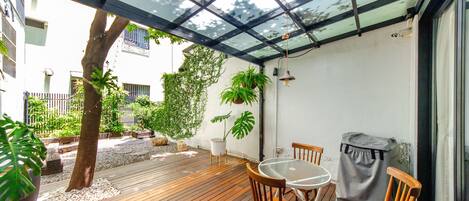A wooden deck with a glass ceiling, surrounded by potted plants and a small table with chairs.
