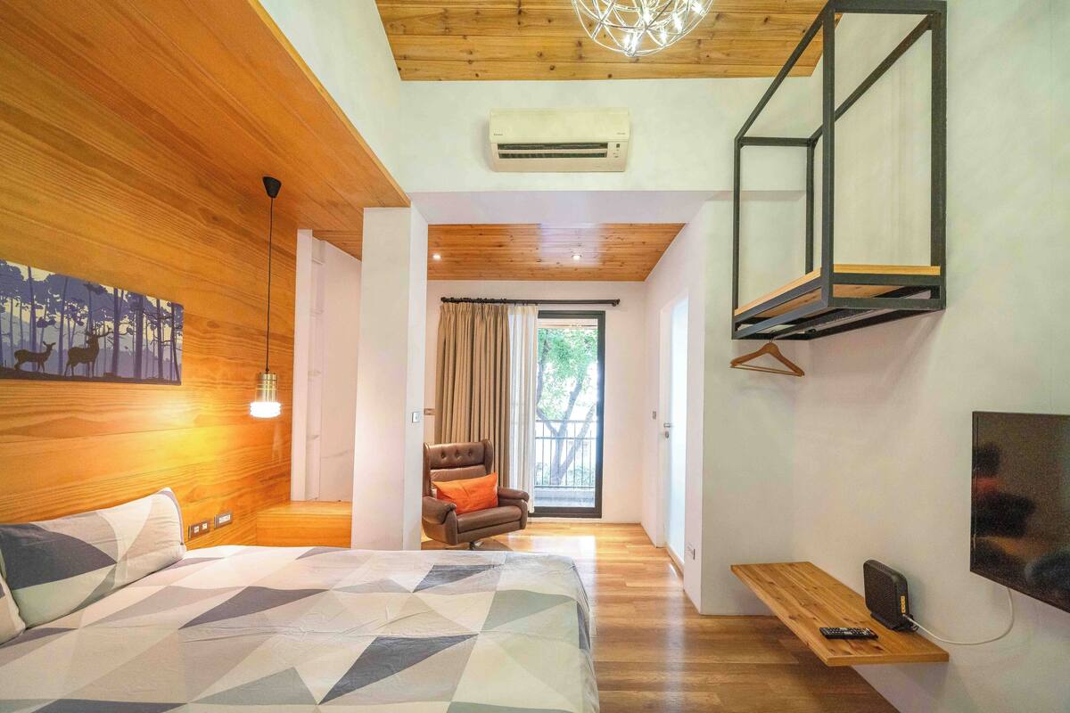 Une chambre avec un plafond en bois, un lit recouvert d’une couette à motifs géométriques, un luminaire suspendu, un téléviseur fixé au mur et une porte coulissante donnant sur un balcon.