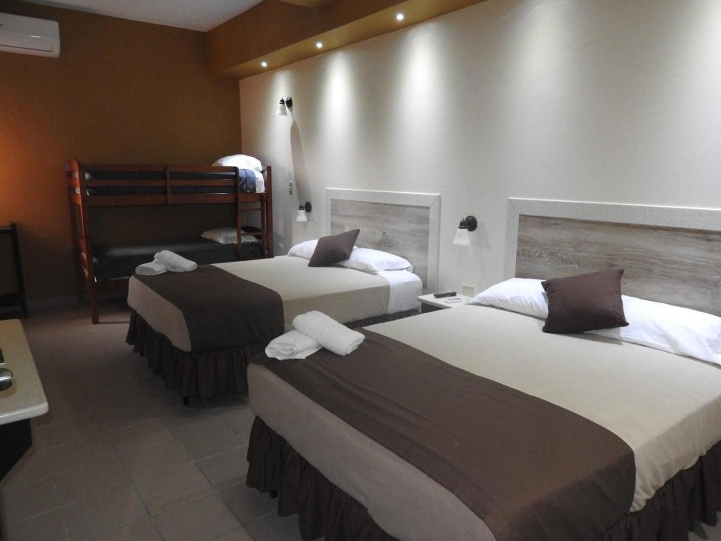 A hotel room with two beds, a bunk bed, a bedside table, and a wall-mounted lamp.