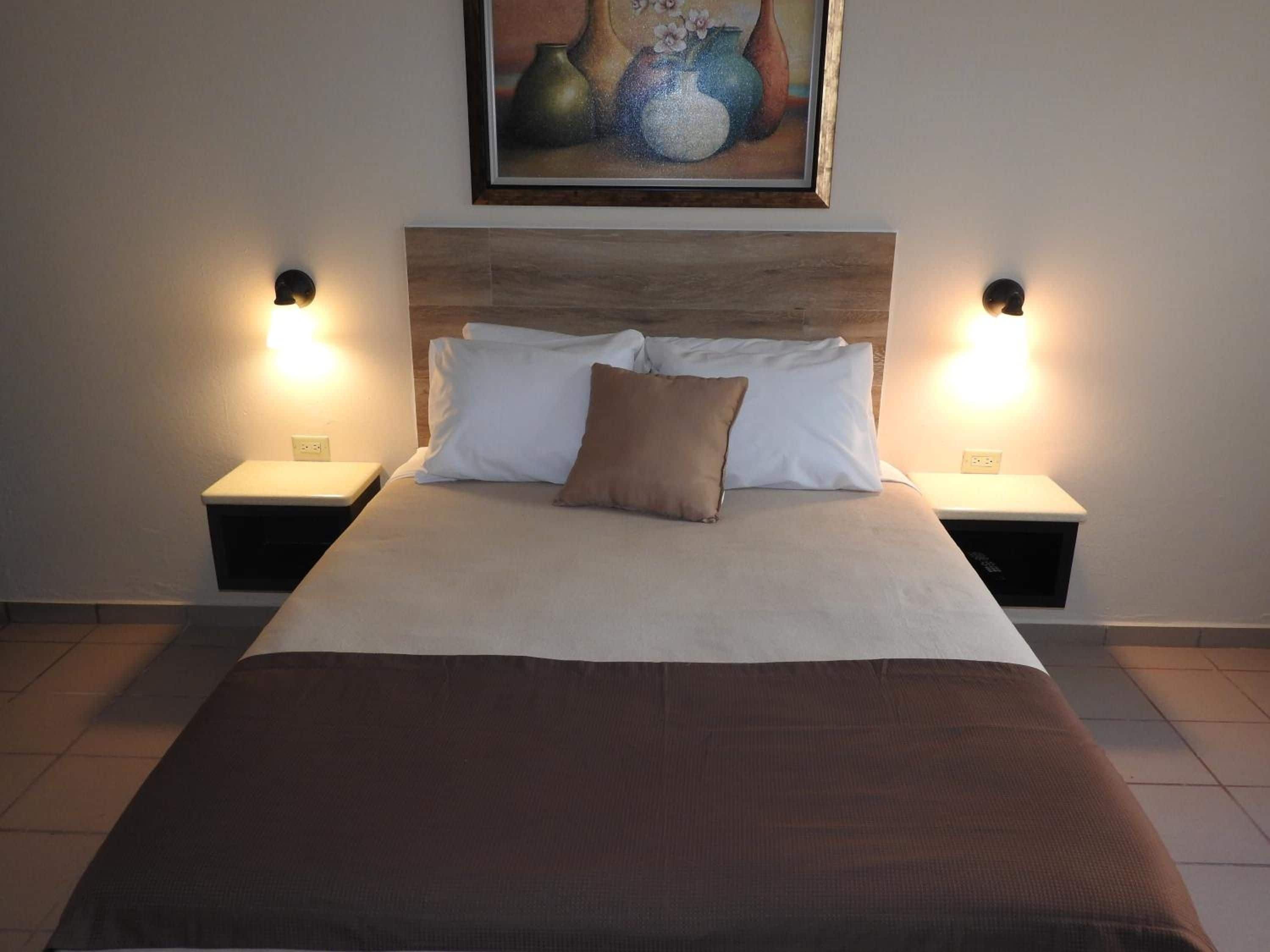 A neatly made bed with a brown blanket, white pillows, and a single brown pillow. There are two bedside tables with lamps on either side of the bed.