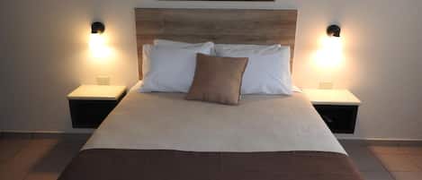 Una cama bien hecha con una manta marrón, almohadas blancas y una sola almohada marrón. Hay dos mesitas de noche con lámparas a cada lado de la cama.