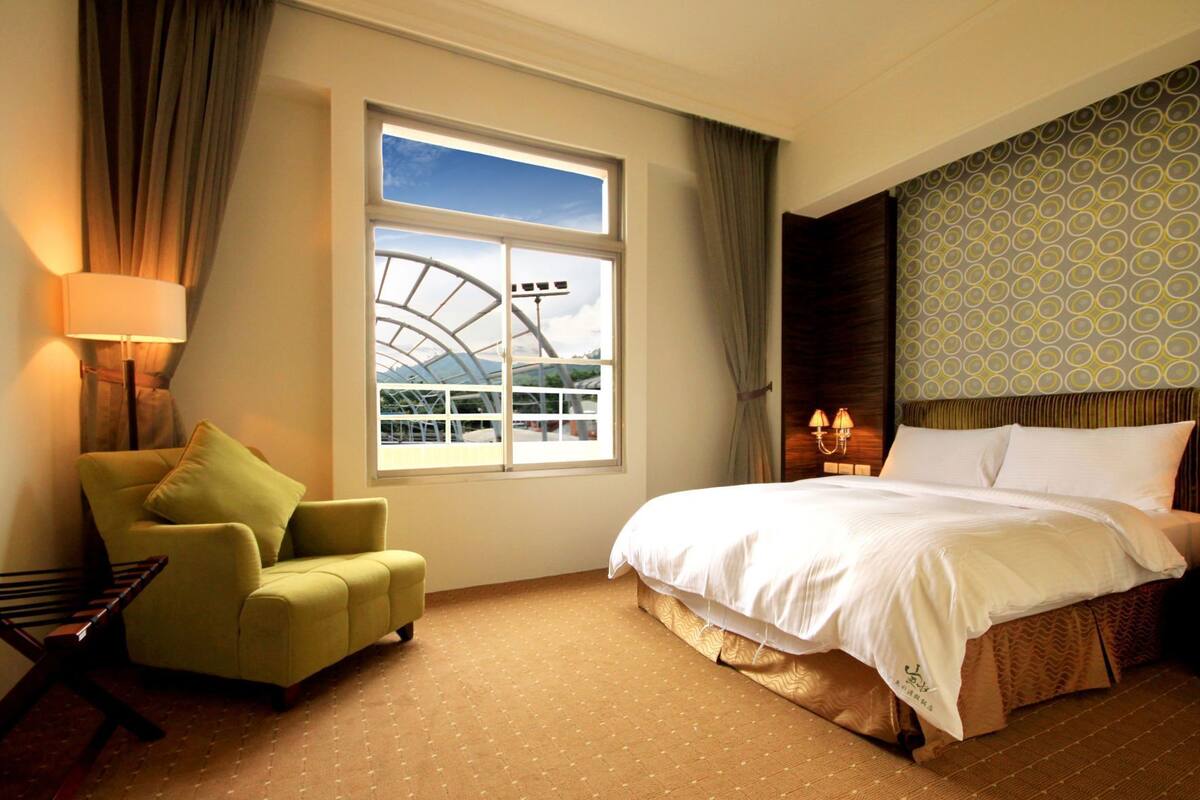 Une chambre d’hôtel avec un lit, une chaise, une lampe, une fenêtre donnant sur un paysage agréable et un mur décoré d’un motif.