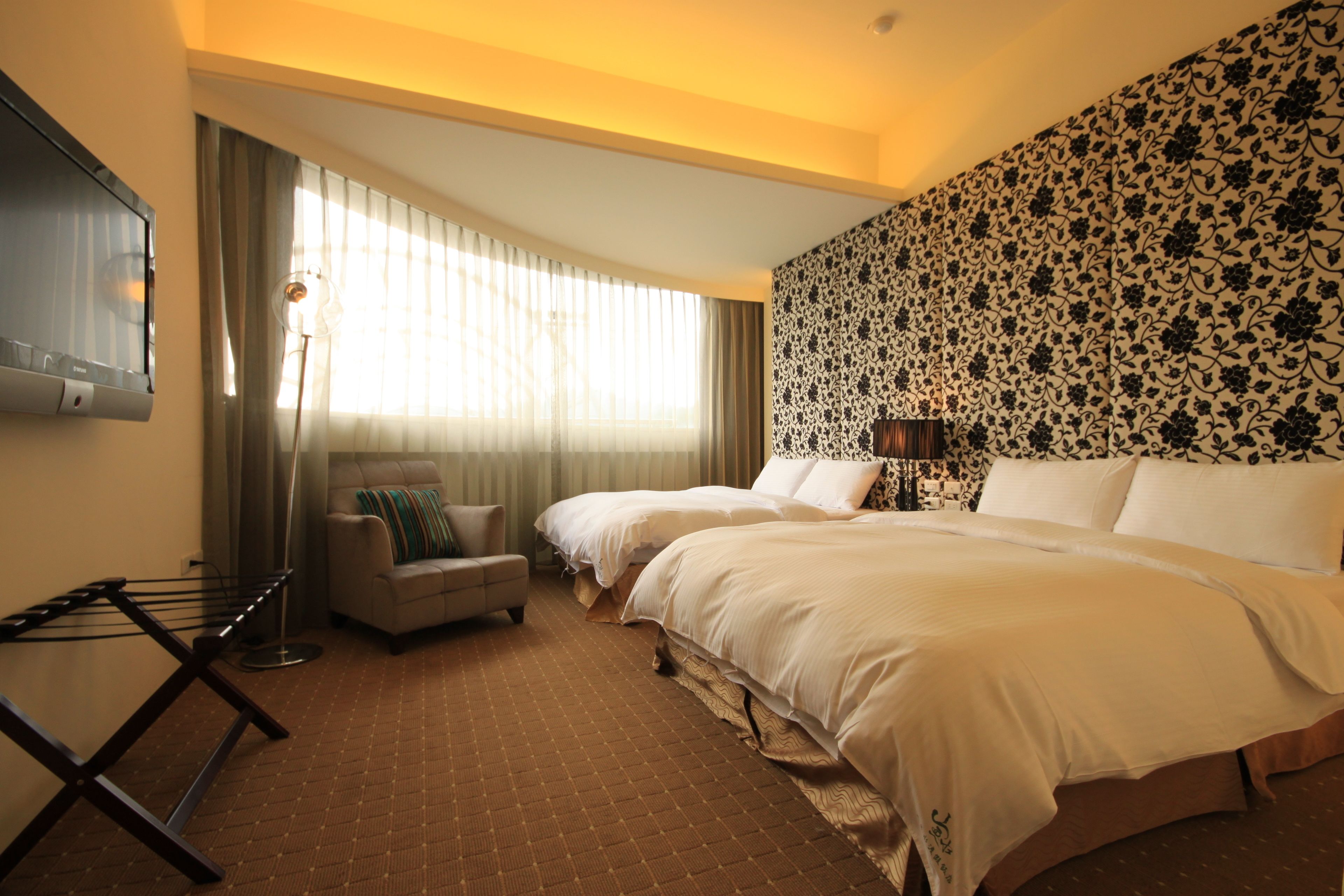 A hotel room with two beds, a flat-screen TV, a seating area with a chair and a table, and a patterned wall.