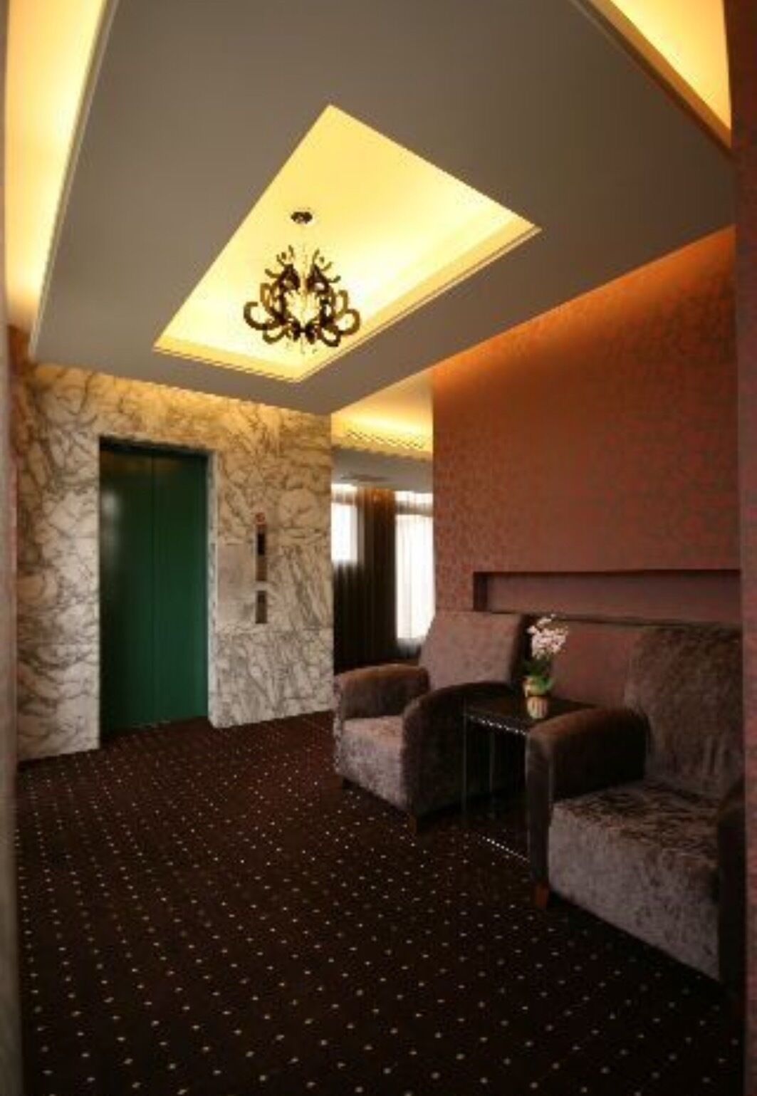 A room with a stone wall, a green door, two armchairs, a small table with a vase, and a patterned carpet.