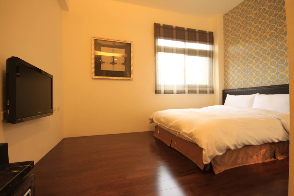 Une chambre à coucher avec un lit, une télévision, une fenêtre avec des rideaux et un mur décoré d’un motif.