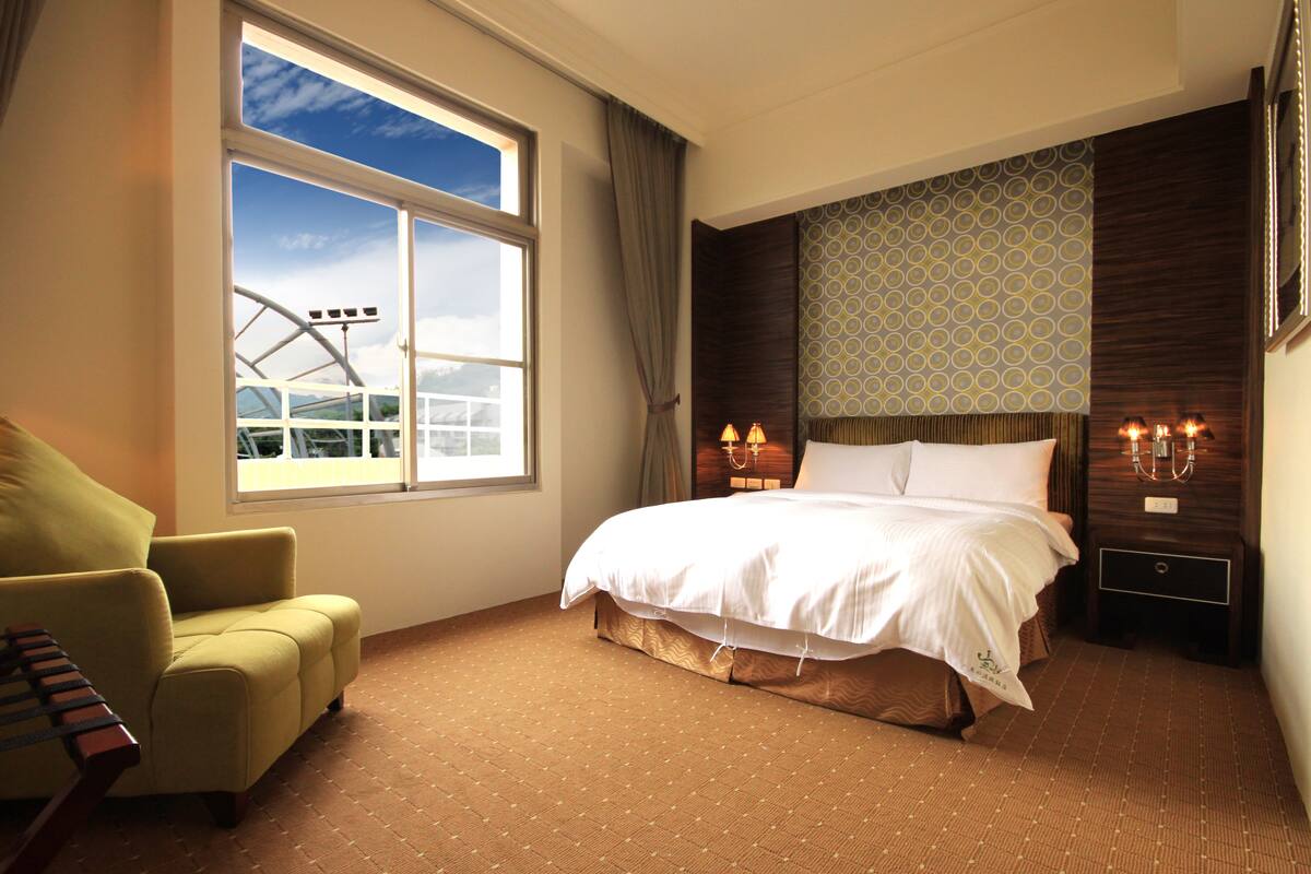 Une chambre d’hôtel avec un grand lit, une chaise, une table de chevet avec des lampes et une fenêtre donnant sur un bâtiment.