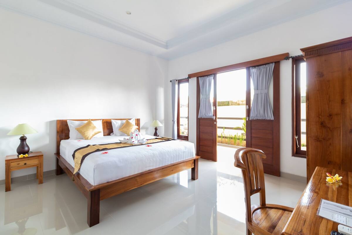 A hotel room with a wooden bed, a wooden chair, a nightstand, and a view of the outdoors through a large window.