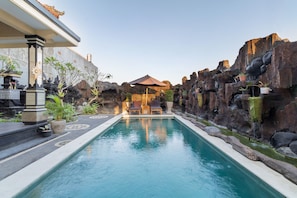 A swimming pool with a stone wall on one side and a building on the other. - Kusuma Guesthouse (Tibubeneng)