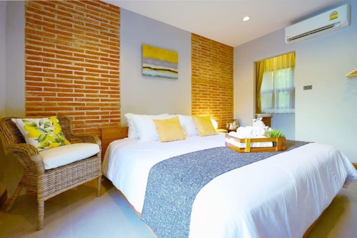 A hotel room with a brick wall, a bed with white bedding, a wicker chair, and a small table with a plant.