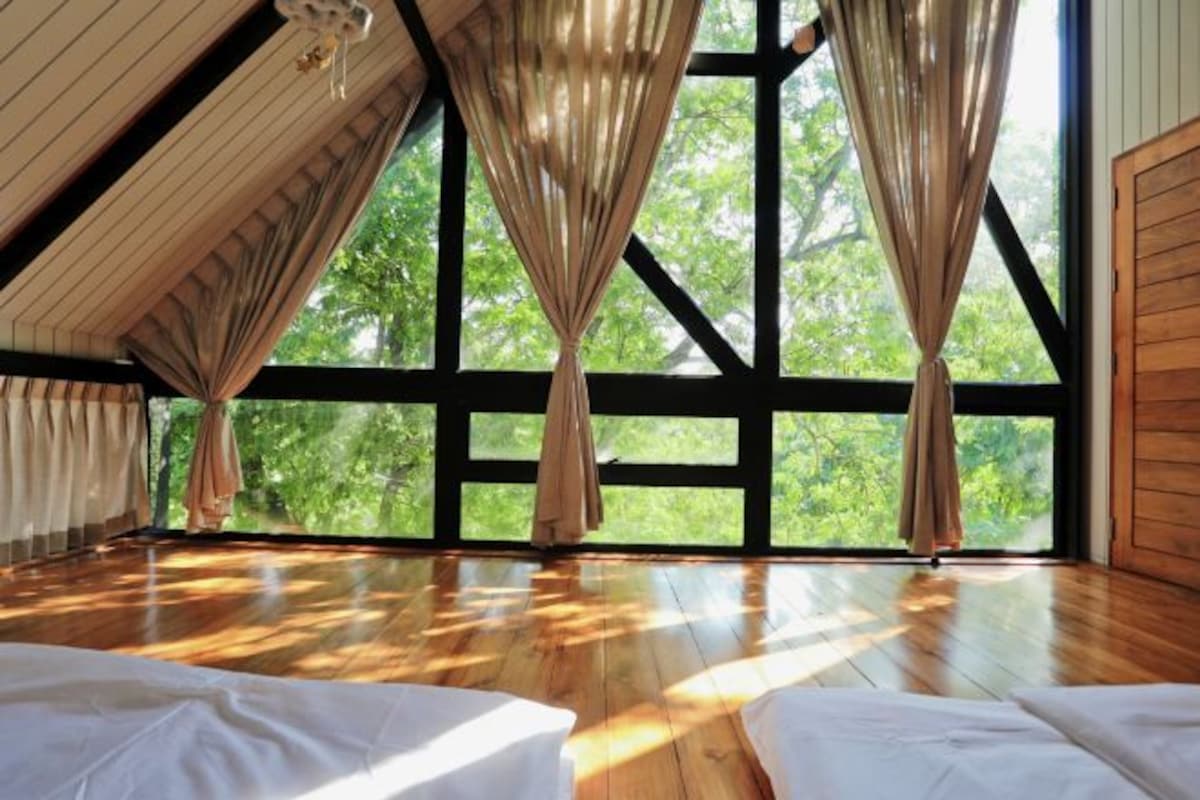 A spacious room with a sloped ceiling, large windows with curtains, and wooden flooring.