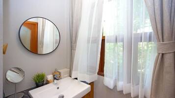 A bathroom with a round mirror, a rectangular mirror, a sink, a potted plant, and a window with sheer curtains.